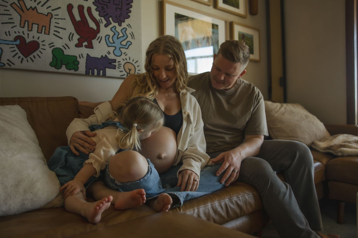 Parents relaxing together on a brown leather couch while bottle feeding their baby at home.
