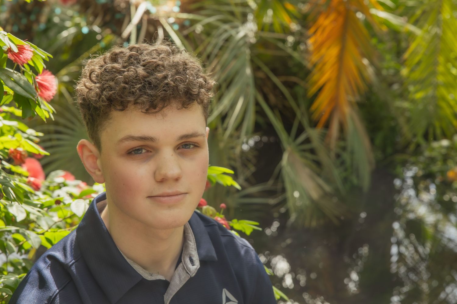 A young person sits outside with lush tropical plants and flowers in the background.