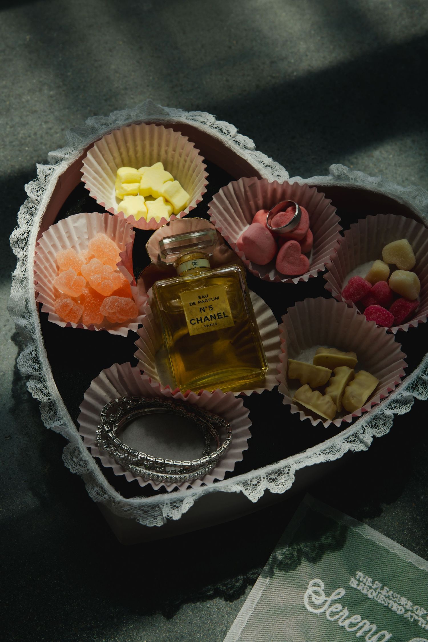A heart-shaped box filled with chocolate candies, sweet treats and a small perfume bottle arranged on dark background.