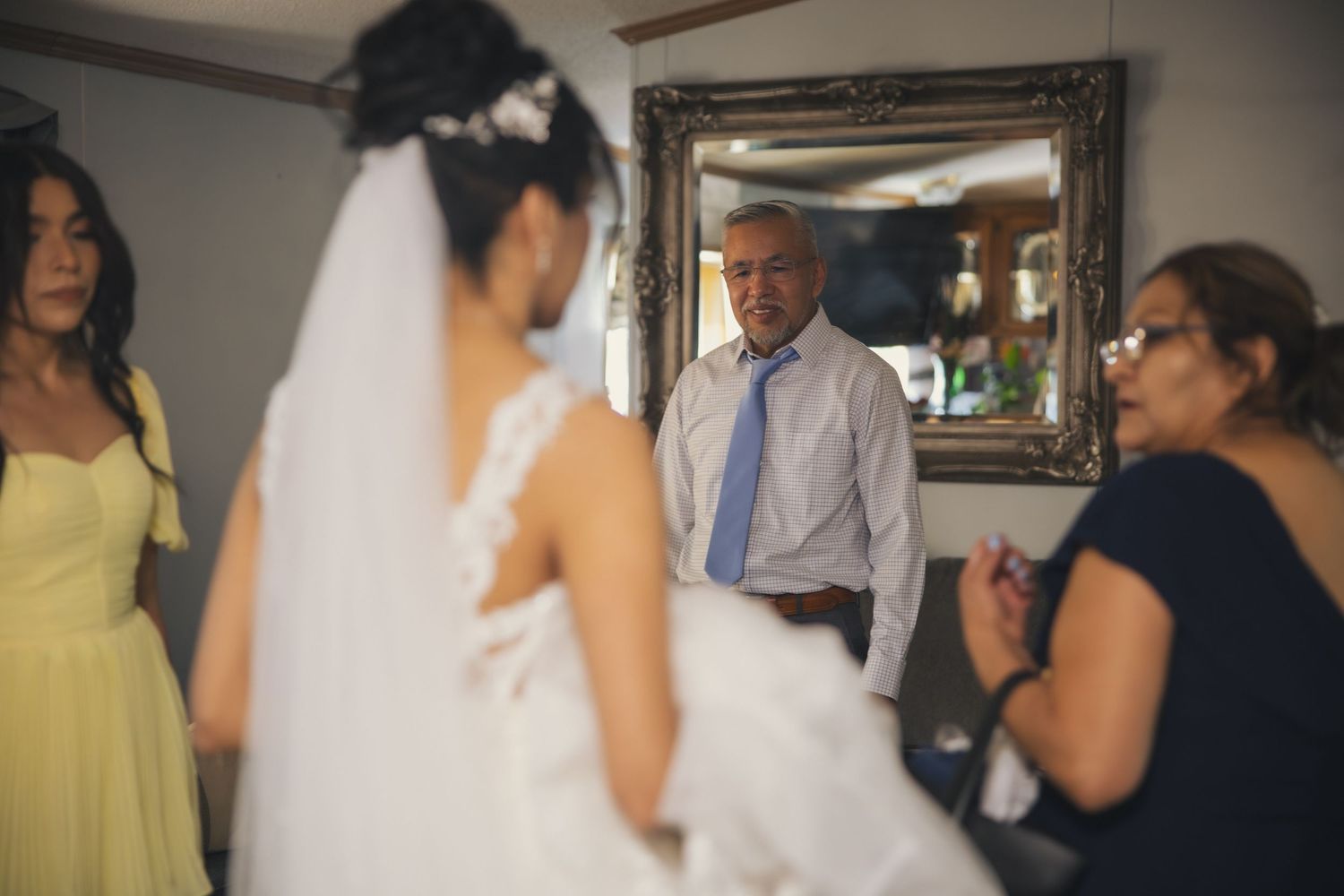 A bride in a white wedding dress is surrounded by family members in a room with a decorative mirror.
