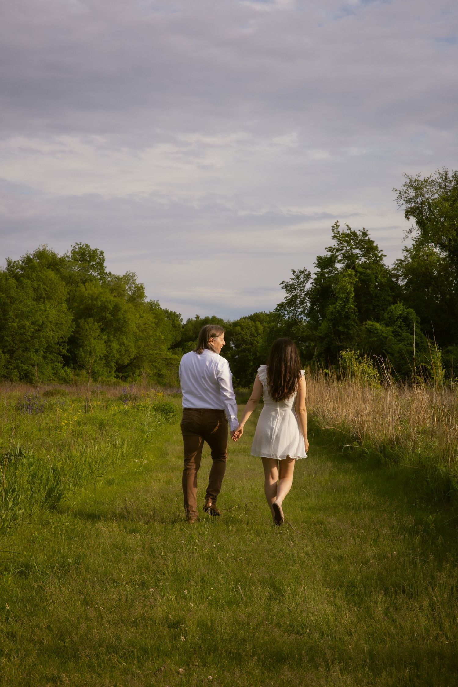 A couple walking together on a grassy trail through a tranquil meadow on a cloudy summer day.