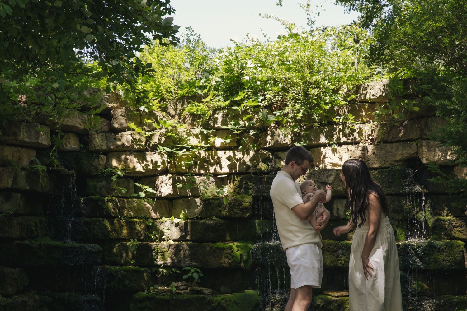 A couple stands embracing against a rustic stone wall backdrop with lush greenery in a garden setting.