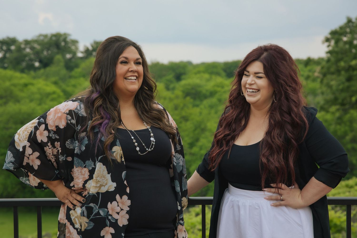Two women share a friendly moment together while standing on a balcony overlooking a green forest landscape.