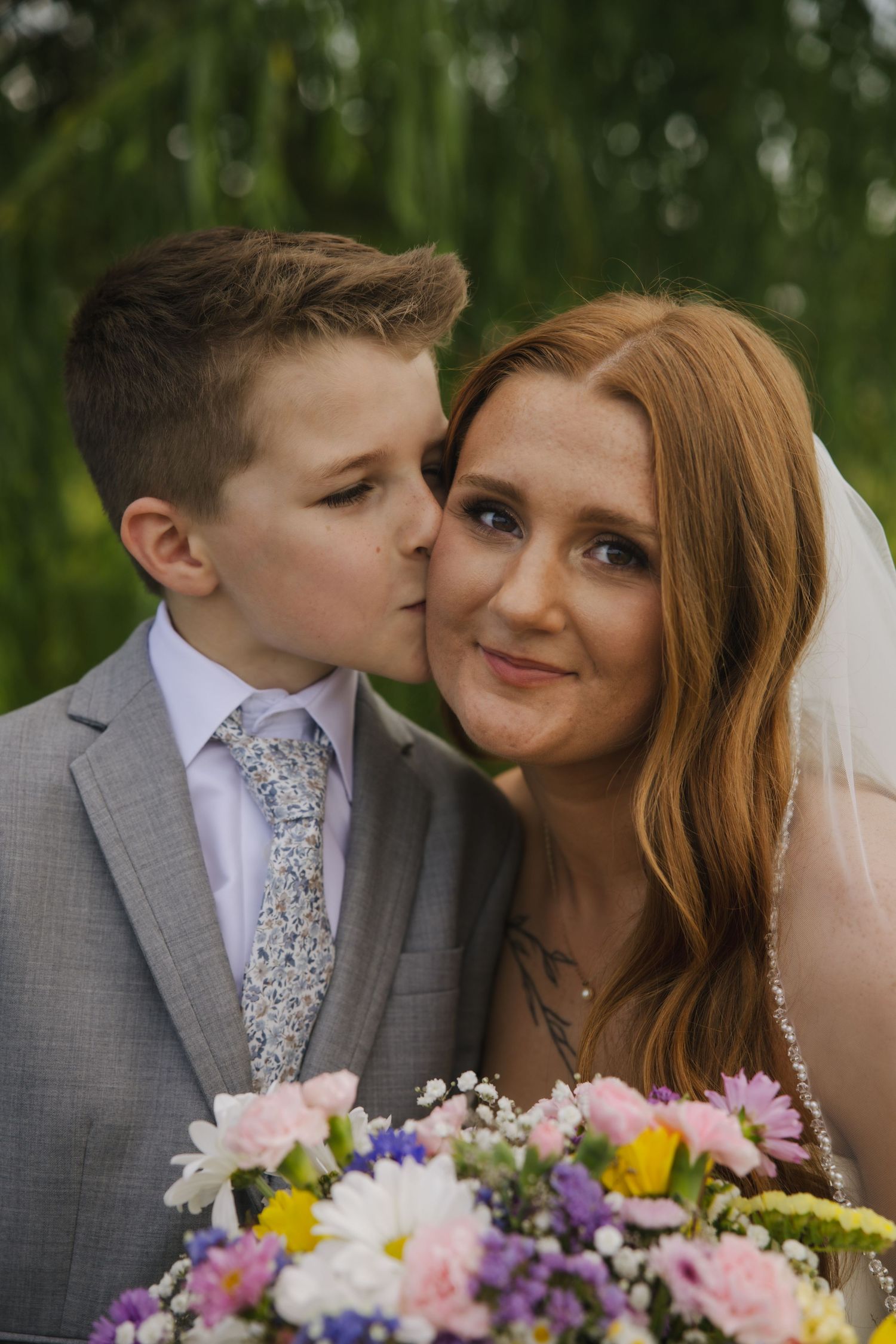 A sweet wedding photo with a gray-suited groom kissing his bride, with colorful flowers in the foreground.