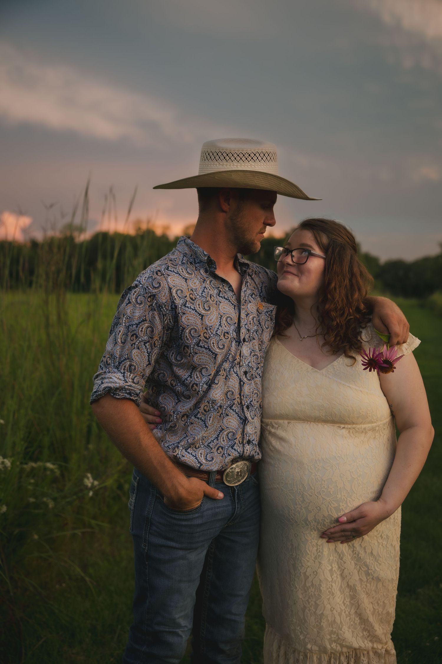 A couple shares an intimate moment in a field at sunset wearing western attire and white lace.