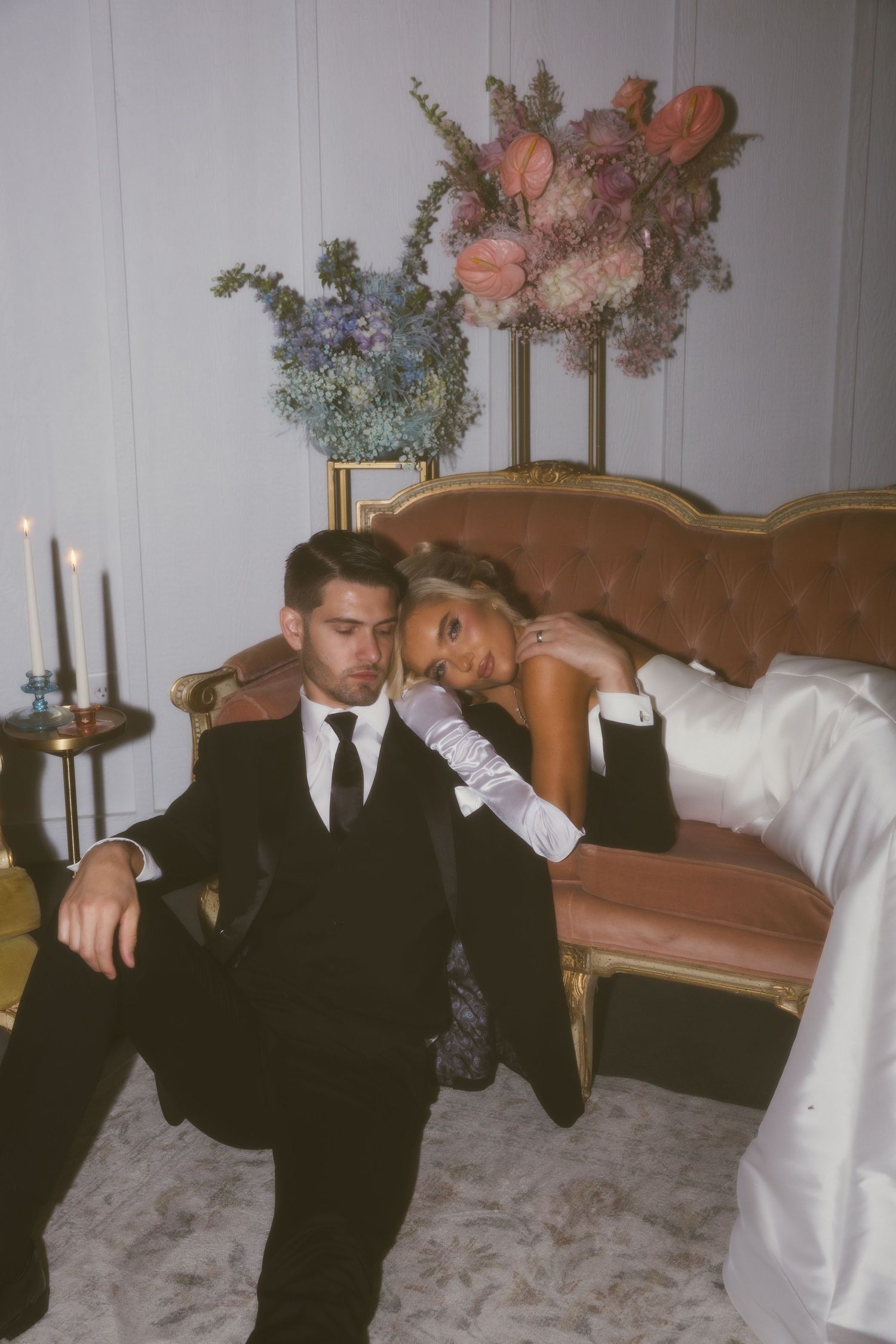 A couple in formal wedding attire sits together on a vintage brown velvet sofa with floral arrangements and candles nearby.