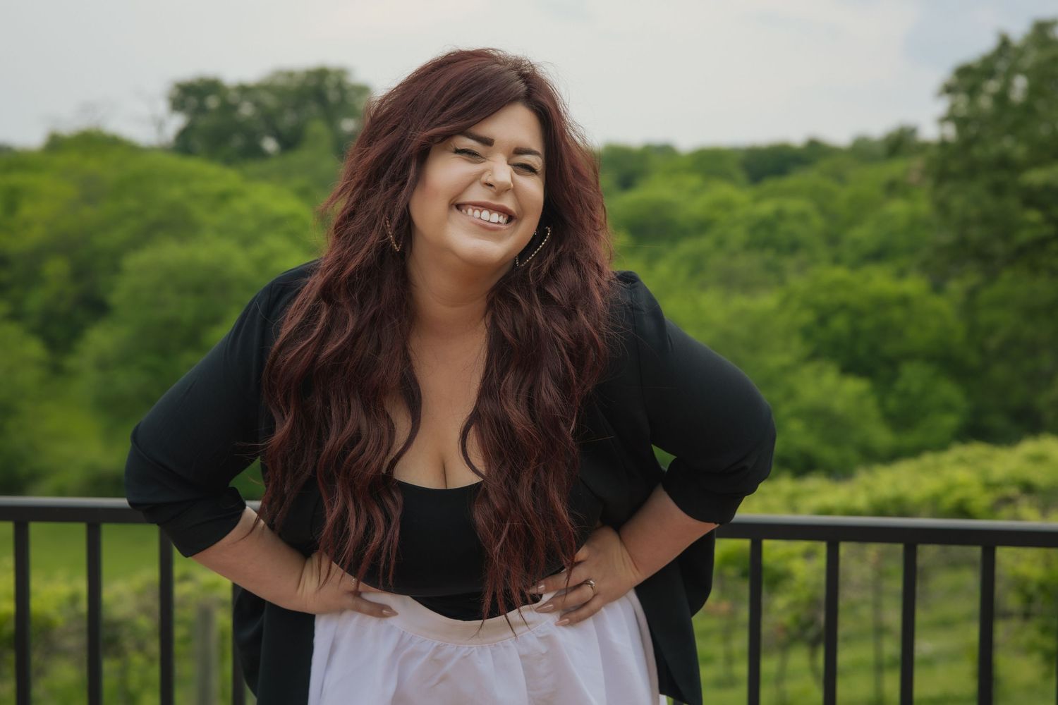 Curvy woman in a black shirt and white skirt smiles warmly while leaning on an outdoor railing with greenery behind.