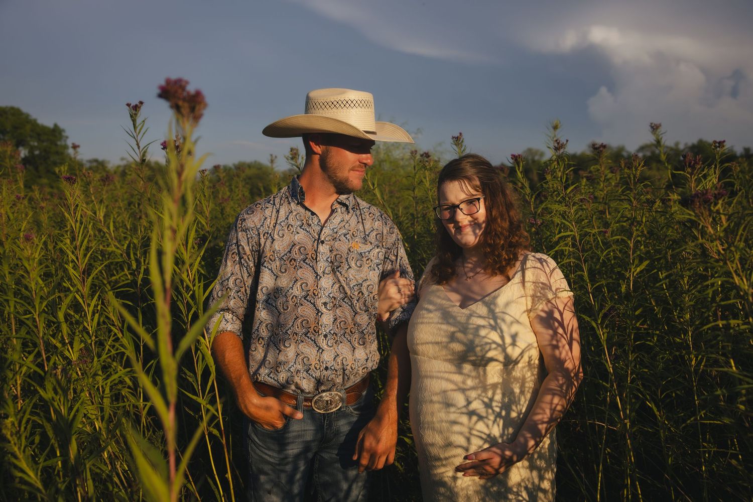 Two people stand together in a tall grass field at sunset wearing rustic country attire.