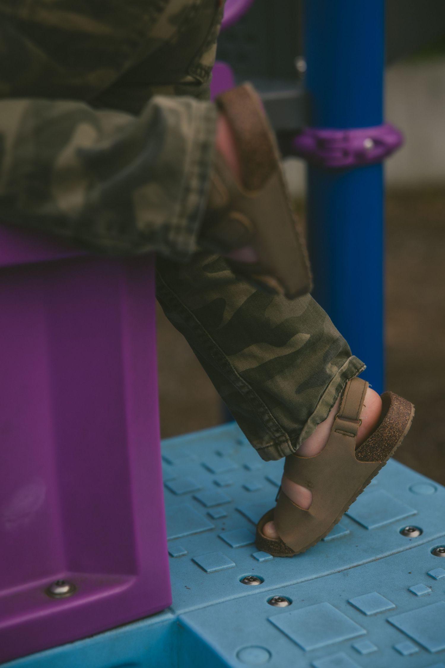 A close-up shot of a child's foot wearing a brown sandal while climbing on blue and purple playground equipment.