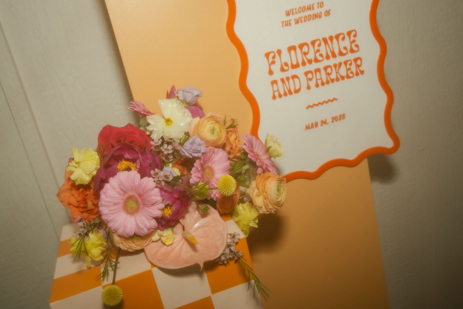 A colorful floral arrangement with pink gerbera daisies and mixed flowers sits beside an orange and white sign.