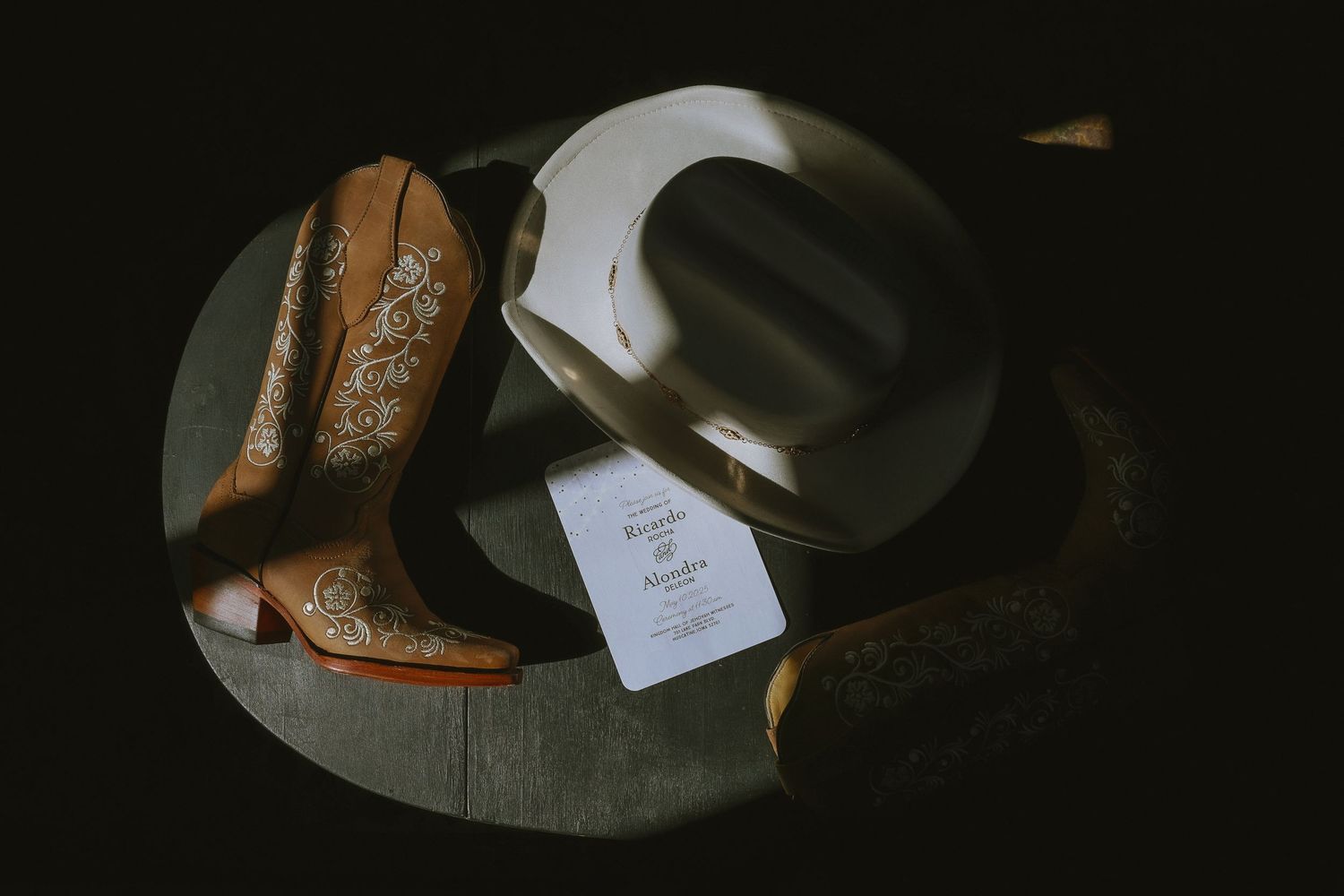 Wedding invitation and hat rest beside brown leather cowboy boots on rustic wooden surface in moody lighting.