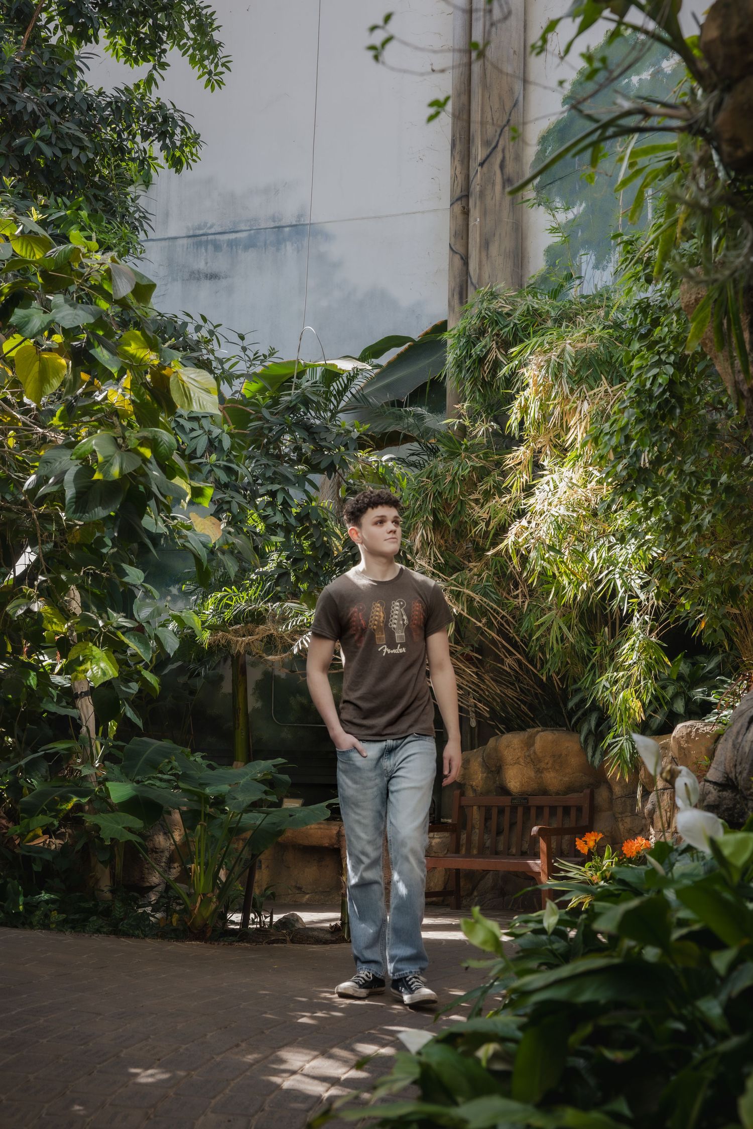 Someone stands in a lush greenhouse garden surrounded by tropical plants and natural sunlight filtering through the foliage.