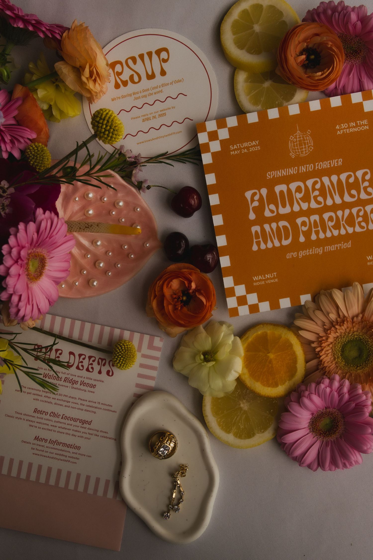 A wedding invitation suite with orange and pink florals, citrus slices, rings, and RSVP card displayed on light surface.