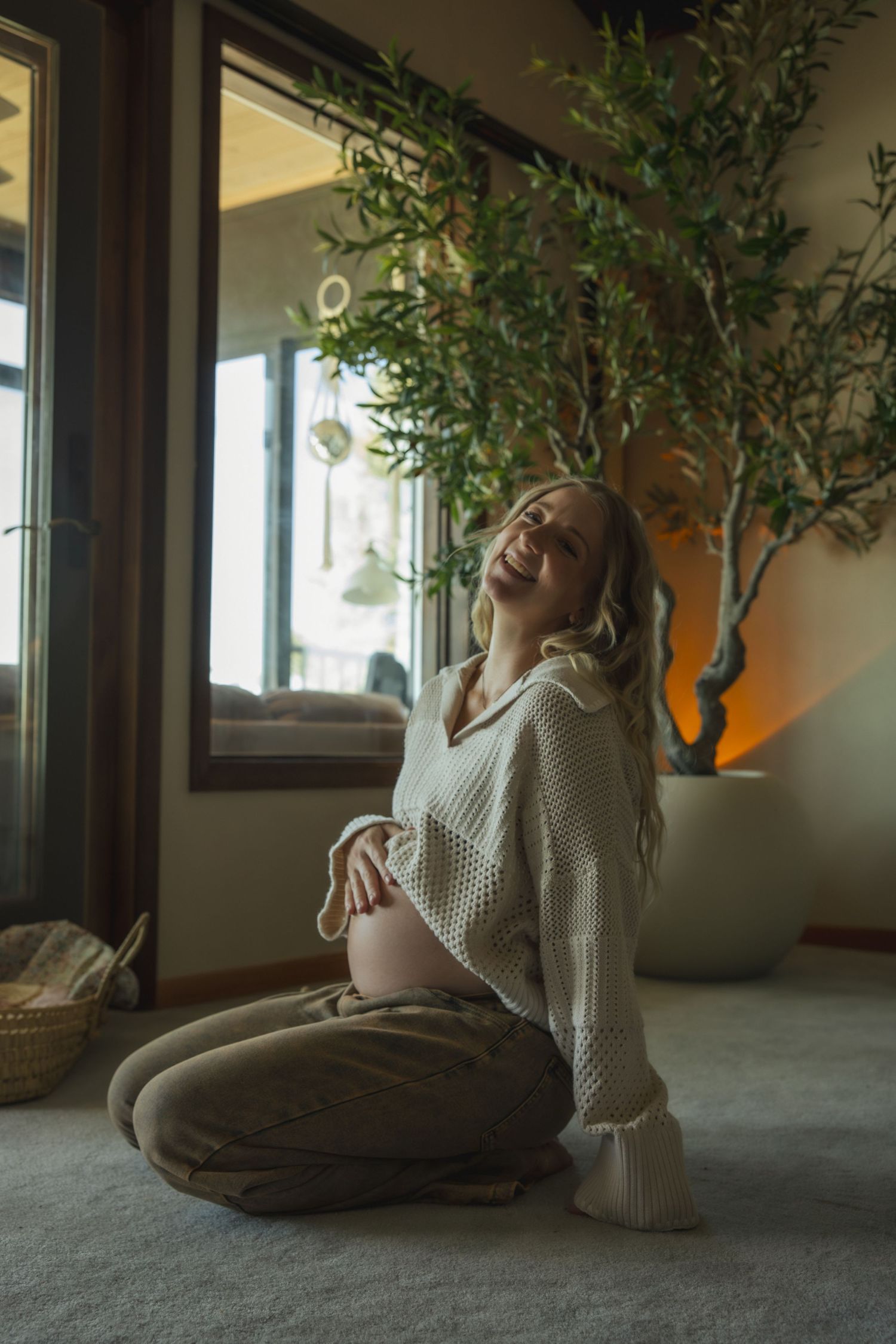 Expectant mother in cozy white sweater sits on floor smiling near indoor plant and window.