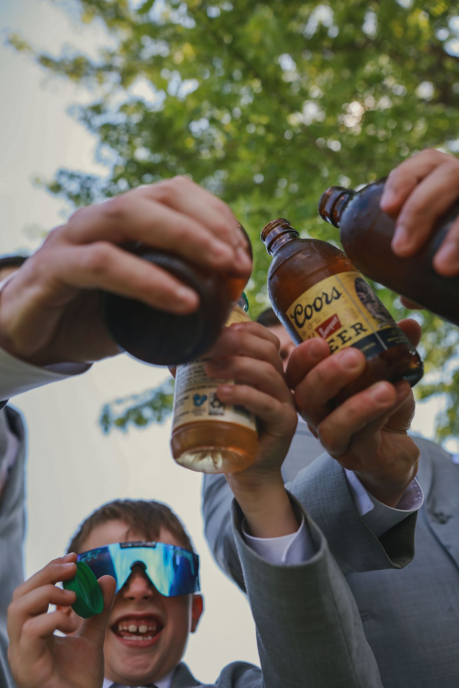 Friends cheering with bottles of Coors beer outdoors on a sunny day against green foliage background.