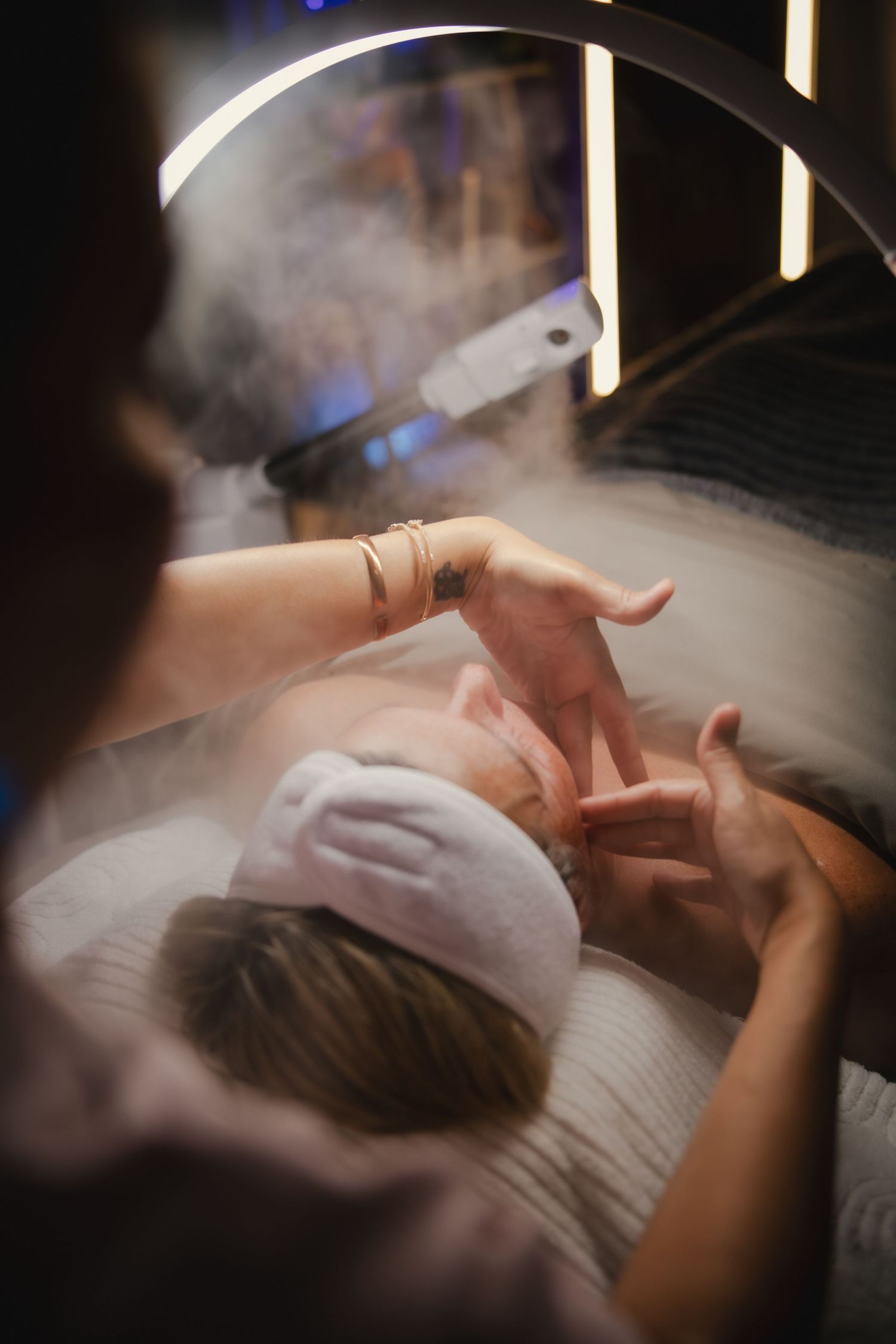 Person lies peacefully in bed covered by white sheets while ethereal smoke or steam drifts through soft lighting.