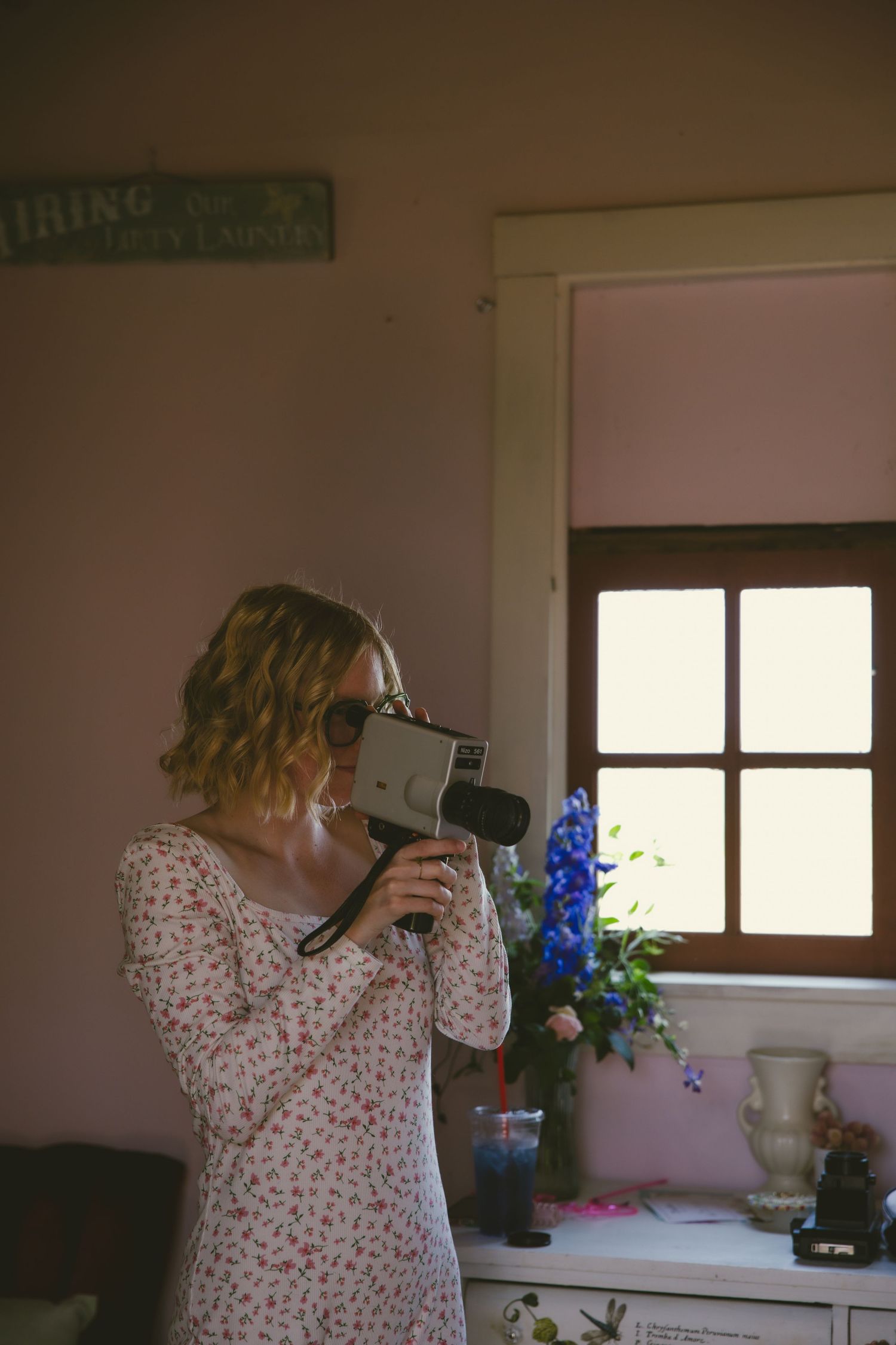 A person wearing a floral dress stands by a window in a dimly lit room with blue flowers in a vase.