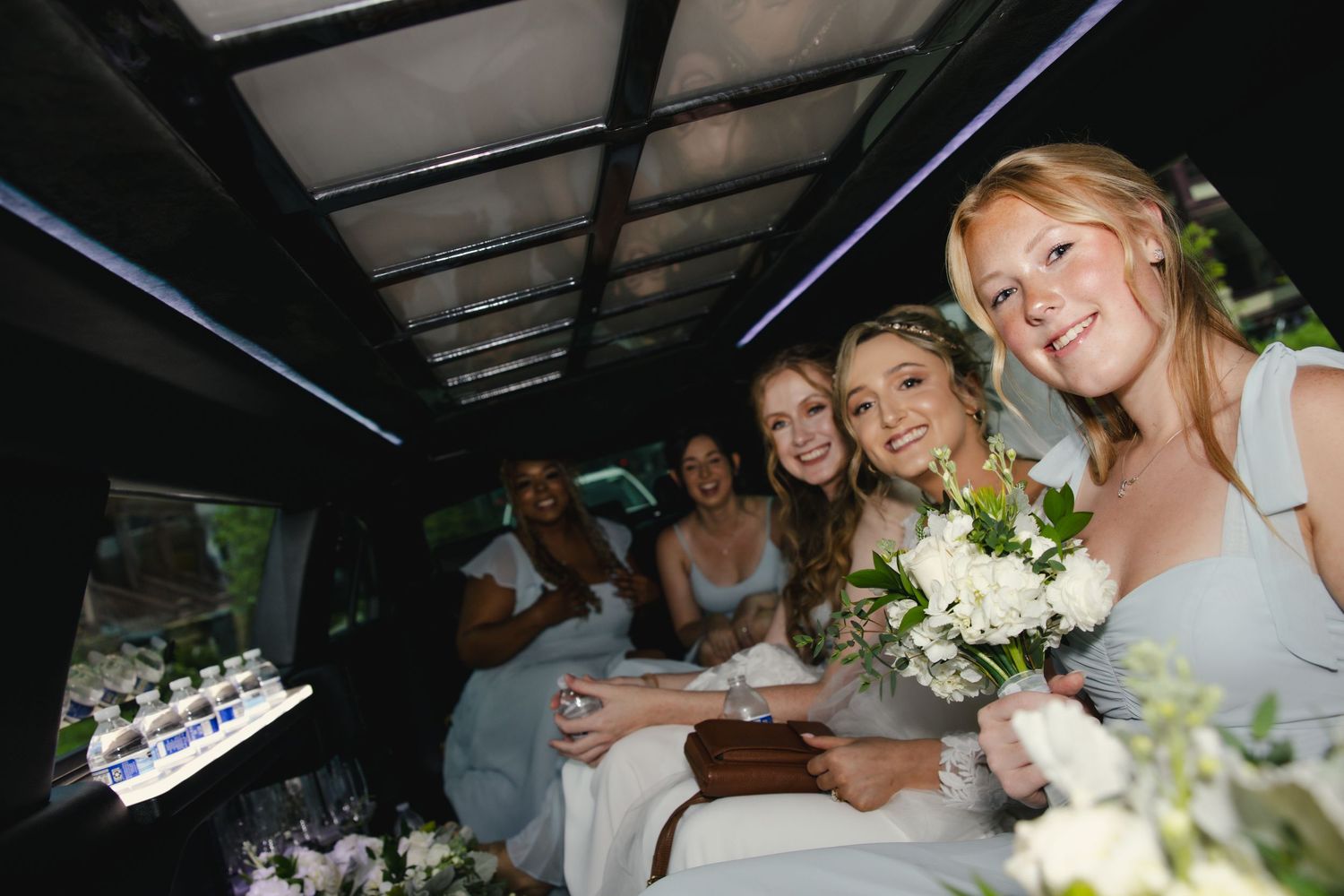A group of people smiling while riding in a limousine with a white floral bouquet.