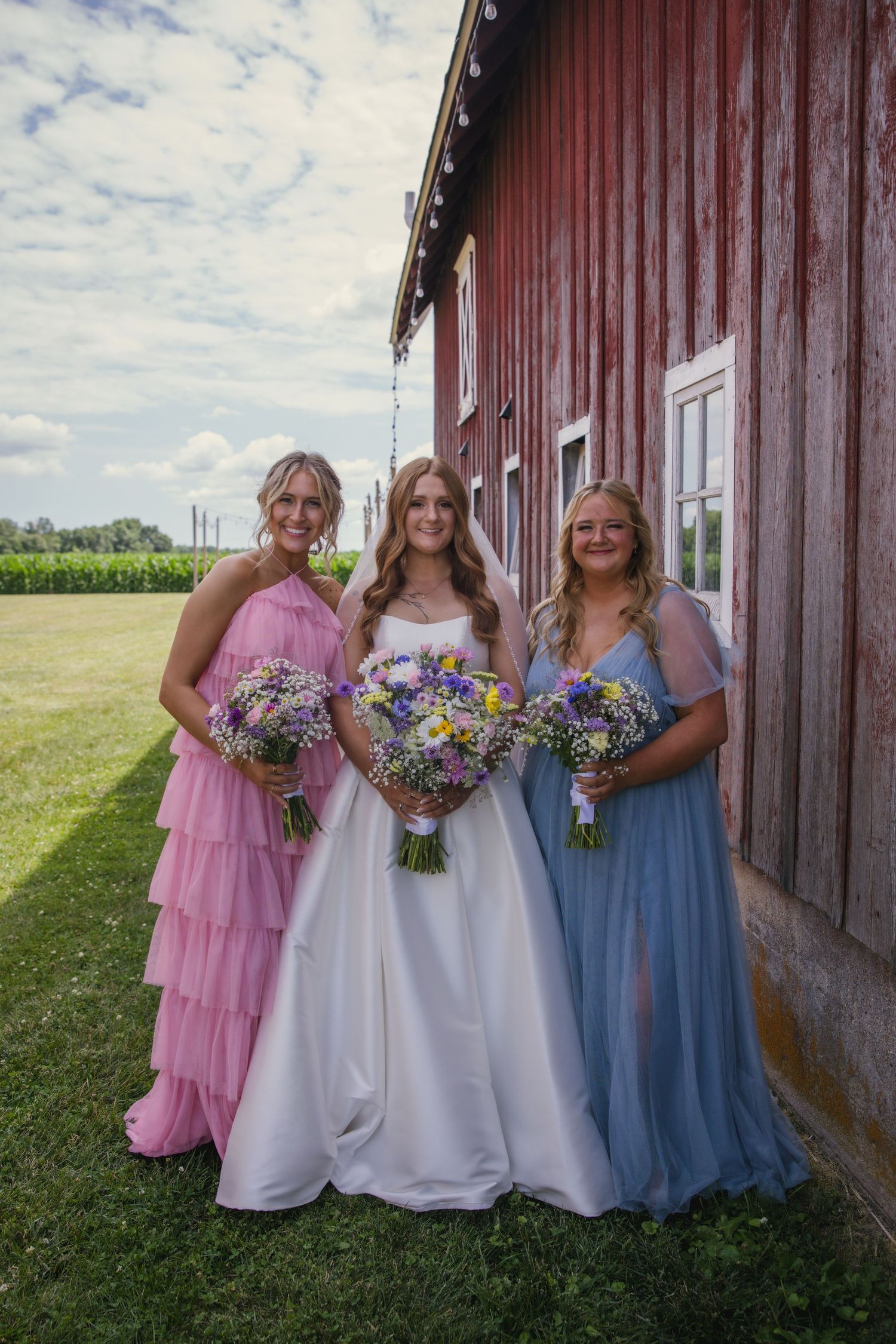 Three people in formal dresses stand by red barn wall holding colorful wildflower bouquets on sunny summer day.