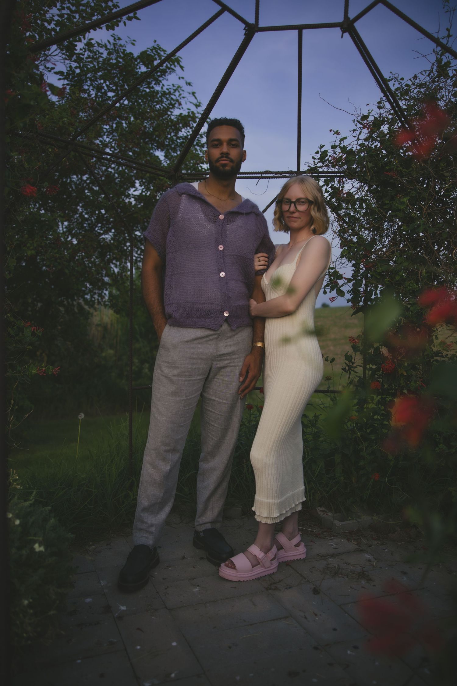 Two people embrace in a romantic dance pose under a garden gazebo surrounded by red flowers at dusk.