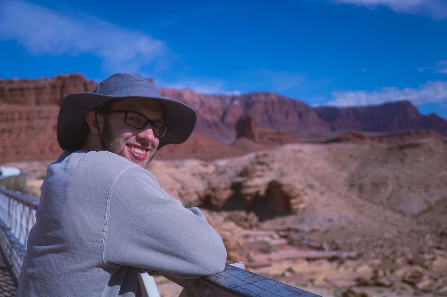 A person in a wide-brimmed hat overlooks red rock desert canyons under blue skies.