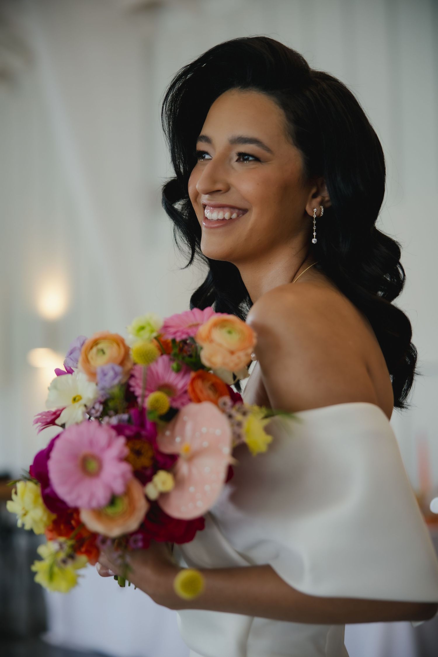 A person in a white off-shoulder top poses with vibrant pink, yellow and orange floral bouquets in a romantic photoshoot.