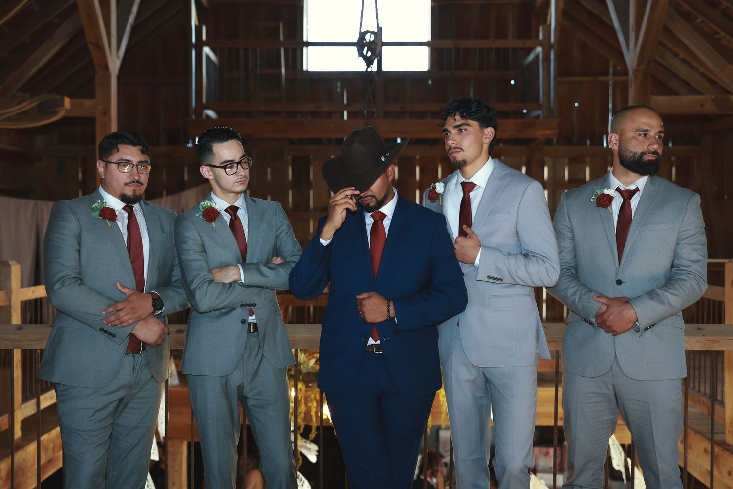 Five groomsmen in coordinated gray and navy suits share a candid moment in a rustic wooden barn venue.
