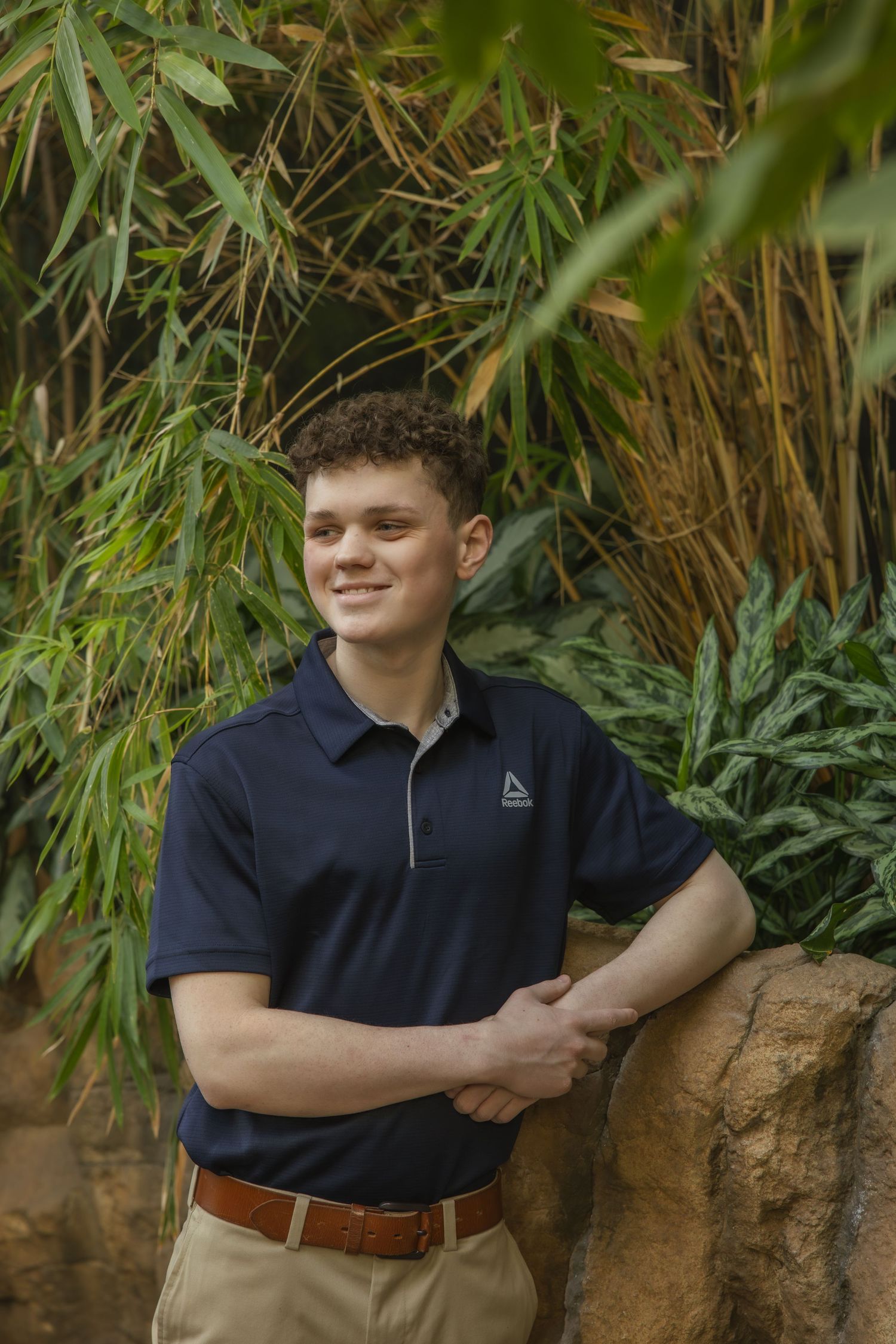Someone wearing a black Reebok polo shirt and khaki pants poses against a natural bamboo and foliage background.