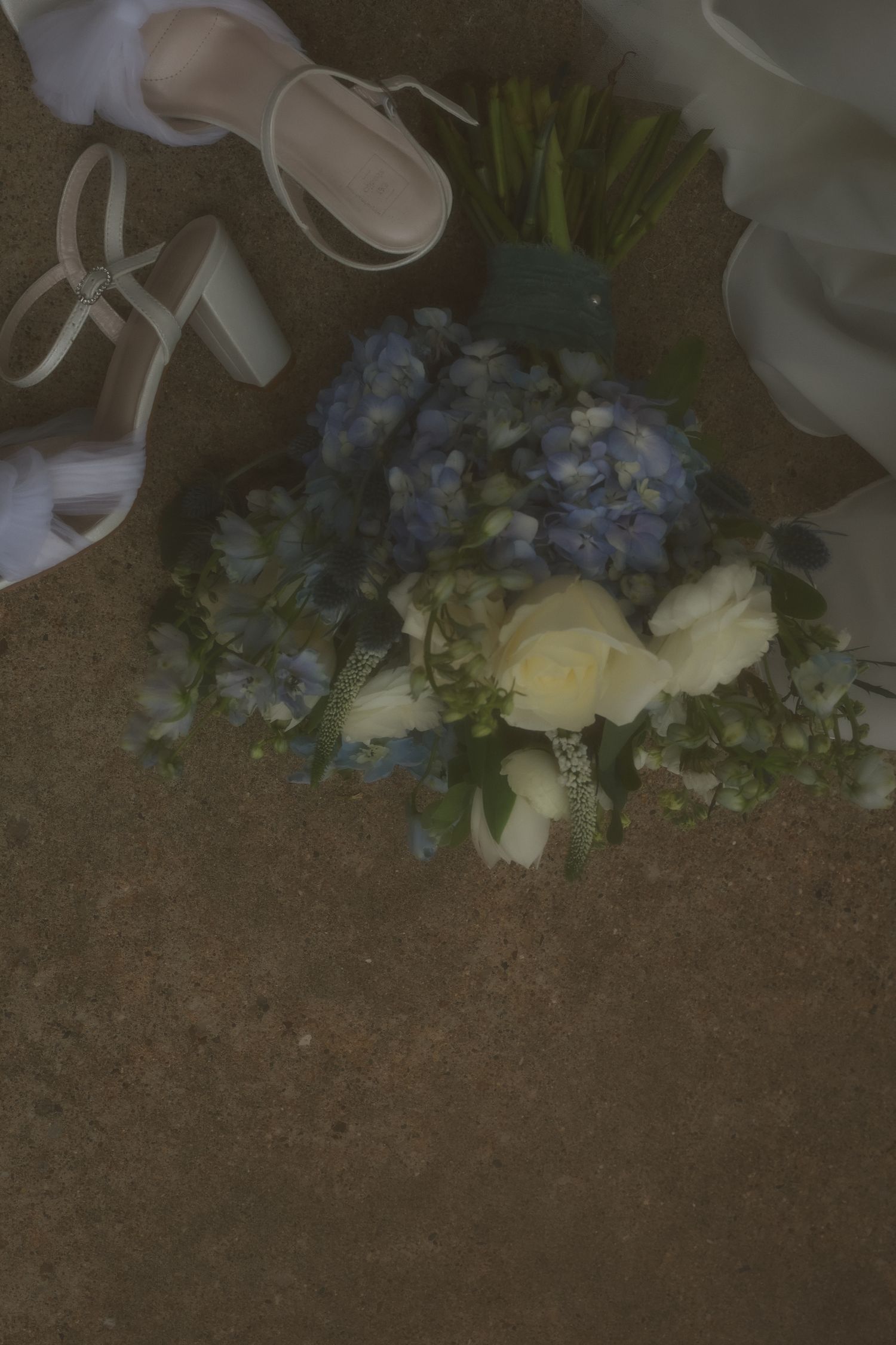 White high heel sandals rest beside a delicate arrangement of blue hydrangeas and cream roses on a textured surface.