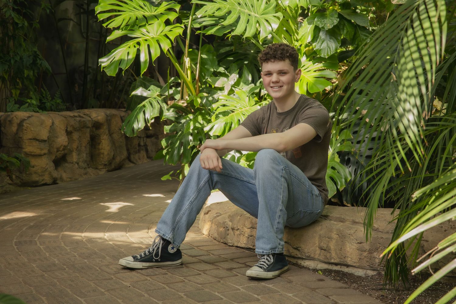 Someone sits casually on stone steps surrounded by lush tropical plants and ferns in a garden setting.