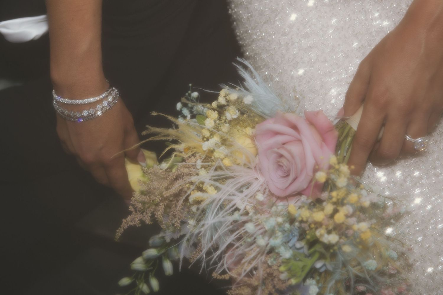 A delicate wedding bouquet featuring pink roses, yellow flowers, and feathery accents against a sparkly white dress.