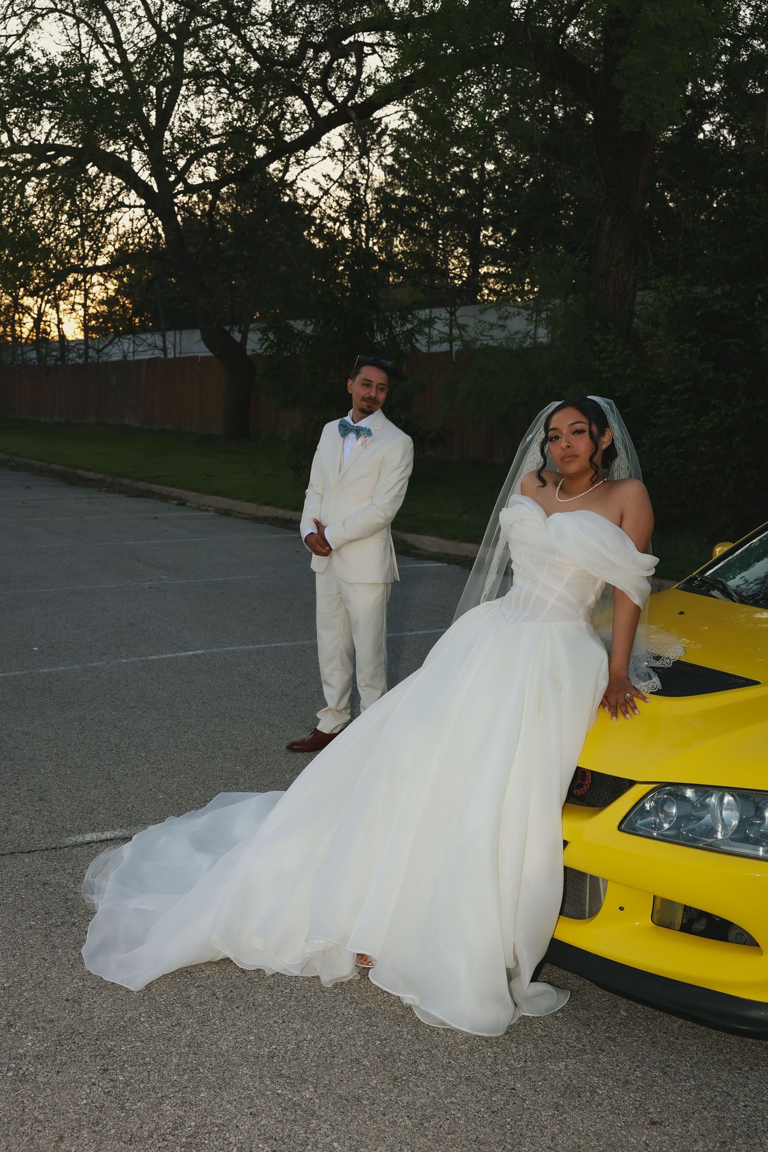A bride in a flowing white dress poses next to a bright yellow sports car at dusk while her partner stands nearby.