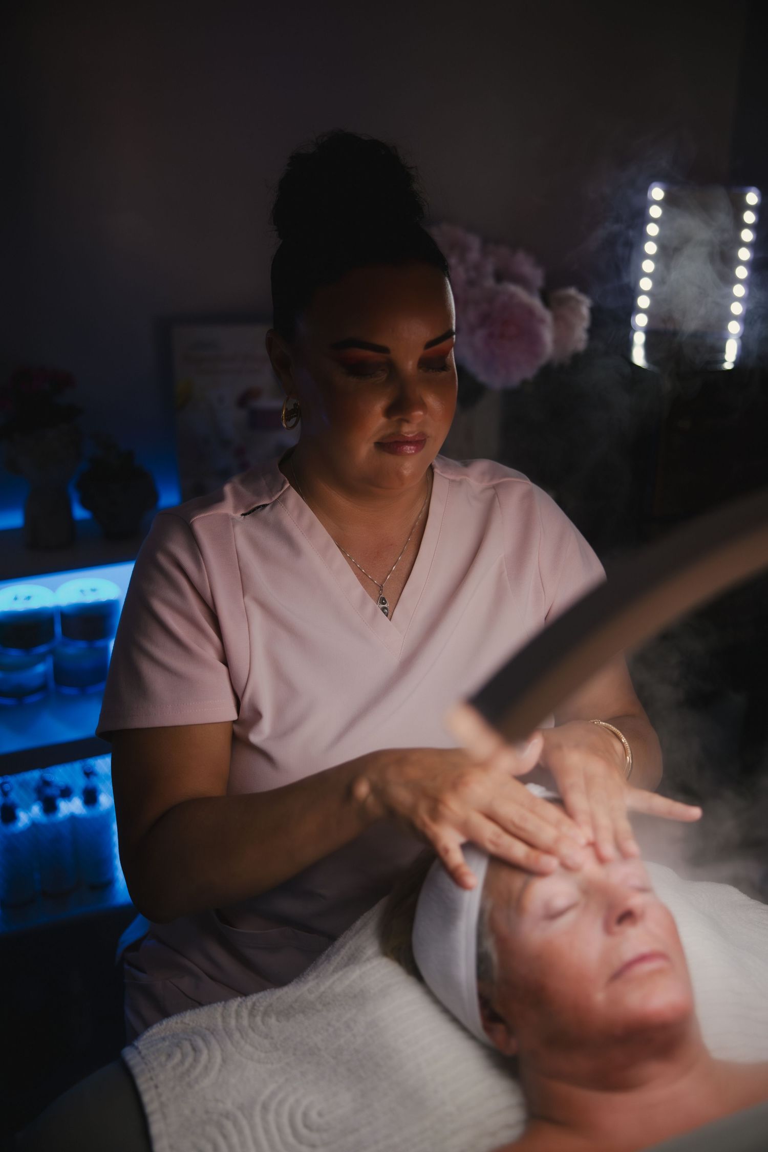 Spa therapist wearing white uniform performs a relaxing facial treatment in a dimly lit room with blue ambient lighting.