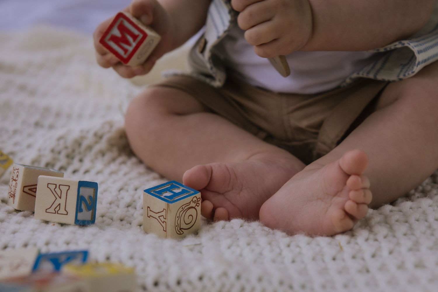 Small feet of a baby sitting on a textured carpet with wooden alphabet blocks and toys scattered around.