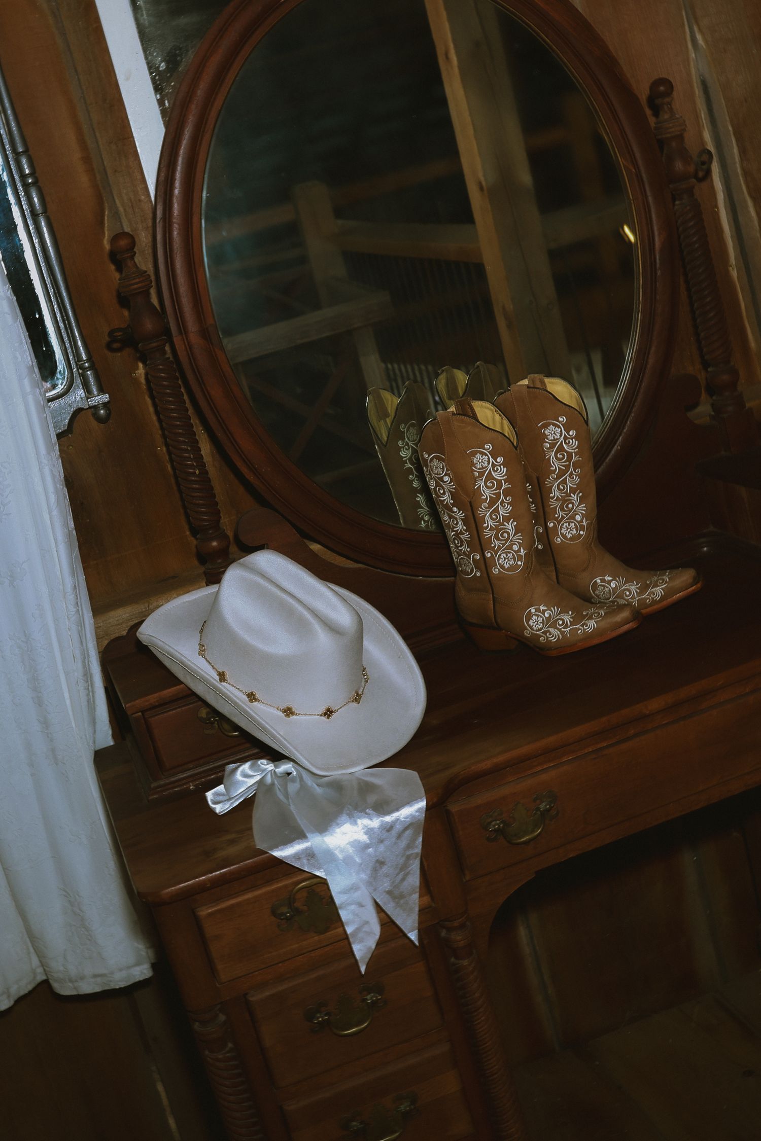 A white cowboy hat and brown cowgirl boots with white embroidery sit atop an antique wooden vanity with an oval mirror.