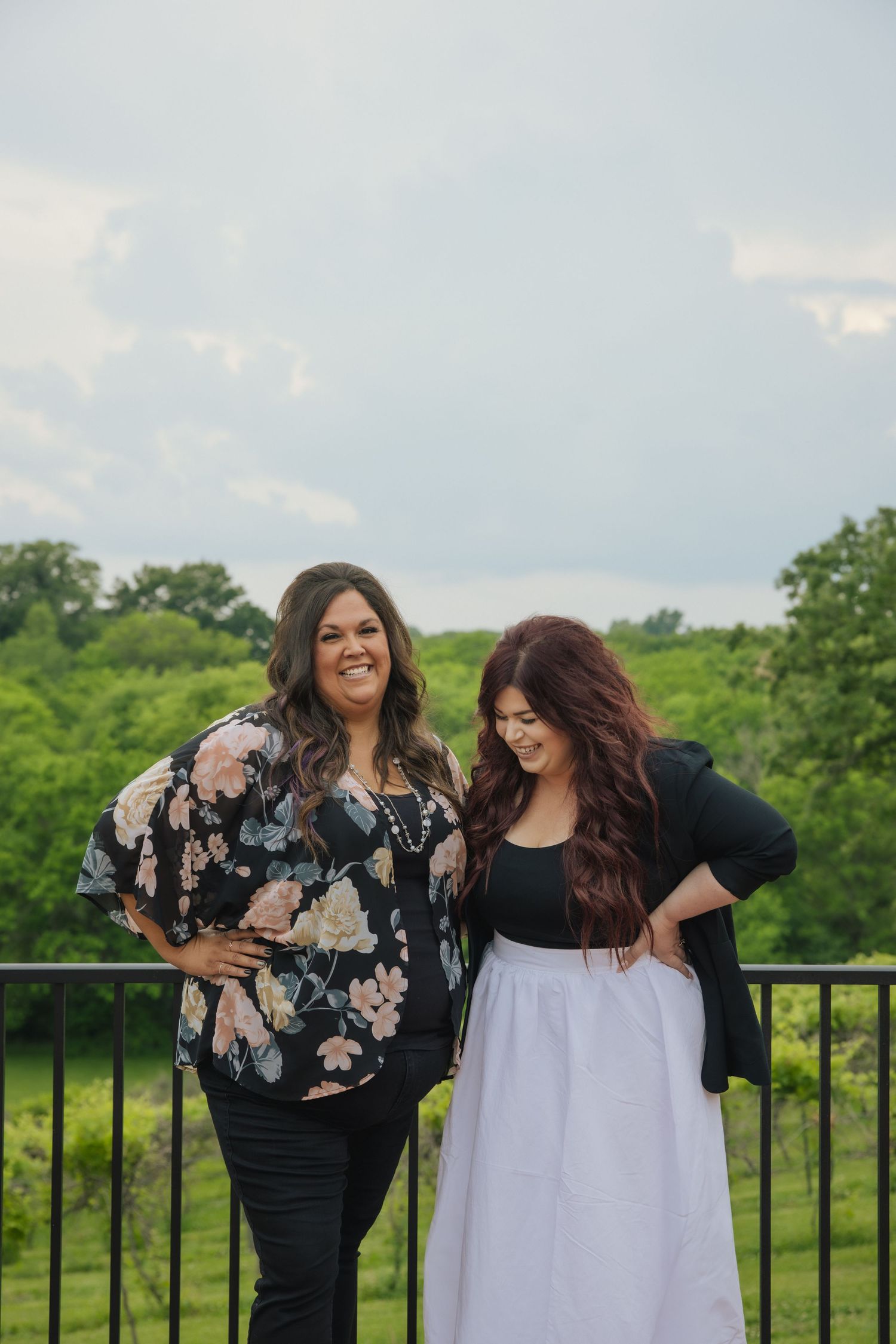 Two women sharing a joyful moment while standing on an outdoor balcony overlooking a lush green landscape.