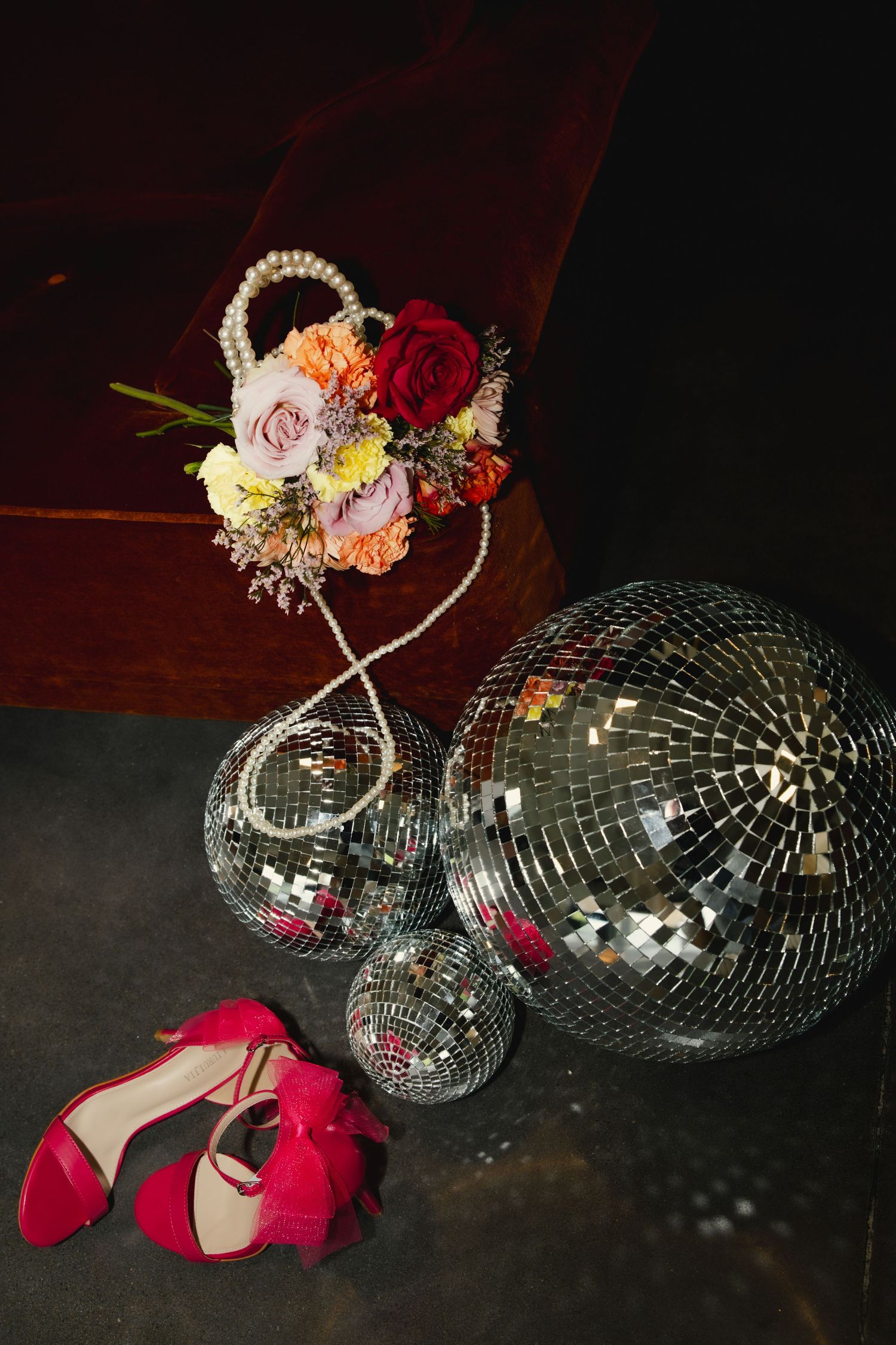 Red heels, disco balls, and a floral arrangement with roses create a glamorous party scene on a dark surface.