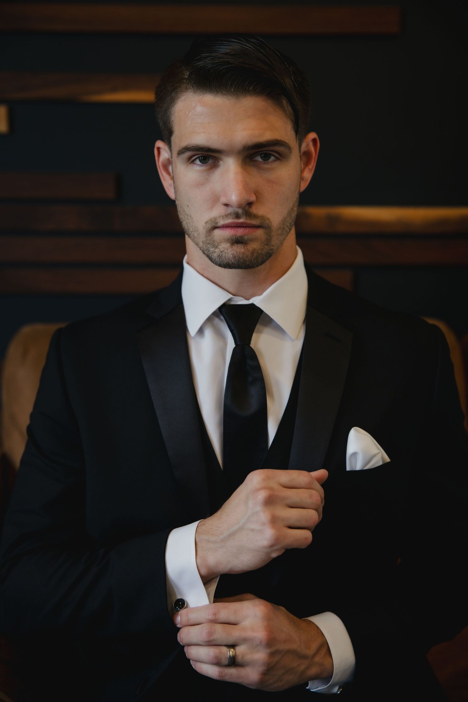 Professional portrait of a person adjusting their cuff in a classic black tuxedo with white pocket square and black tie.