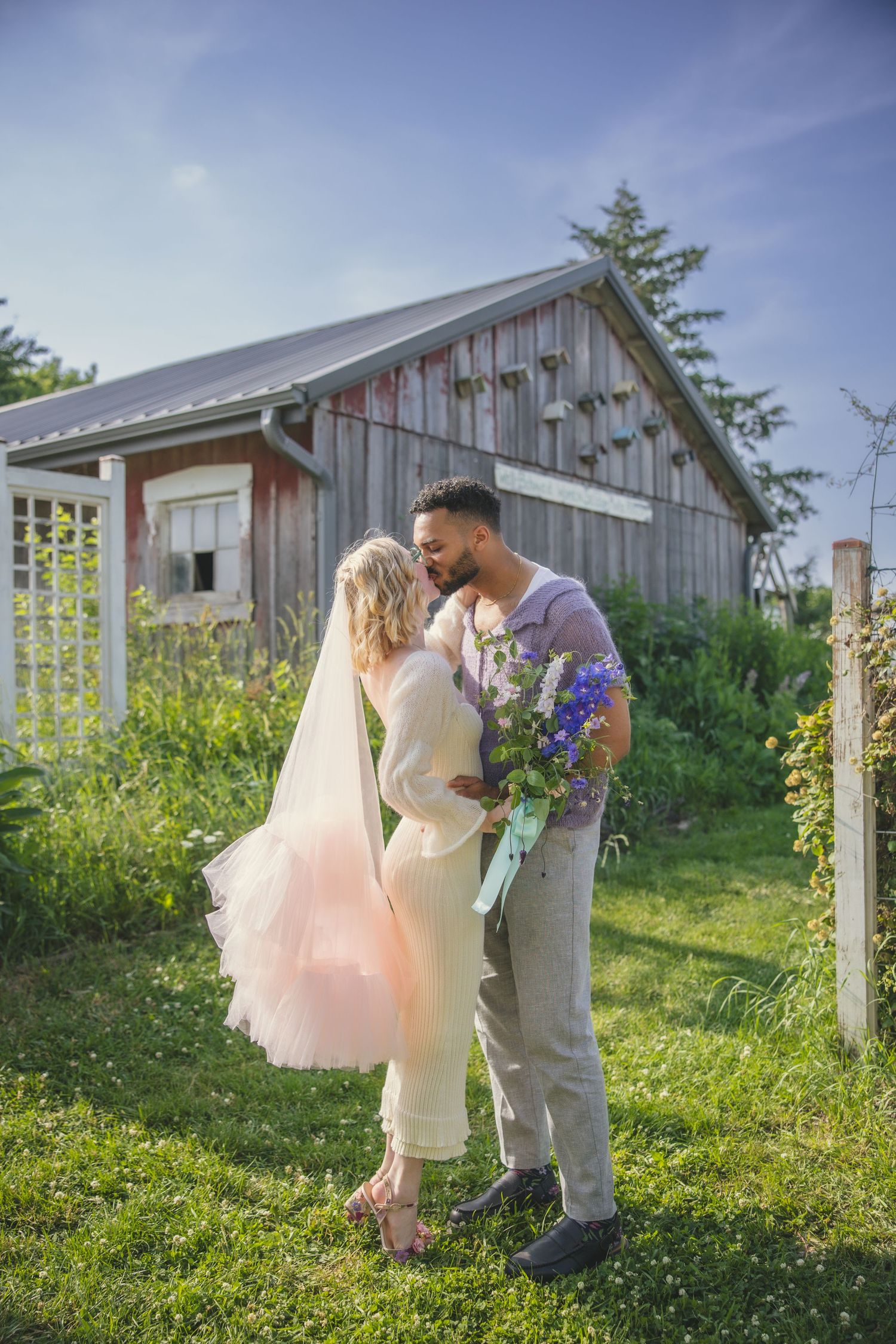 A couple stands together in front of a rustic barn on a sunny day, surrounded by lush greenery and wildflowers.