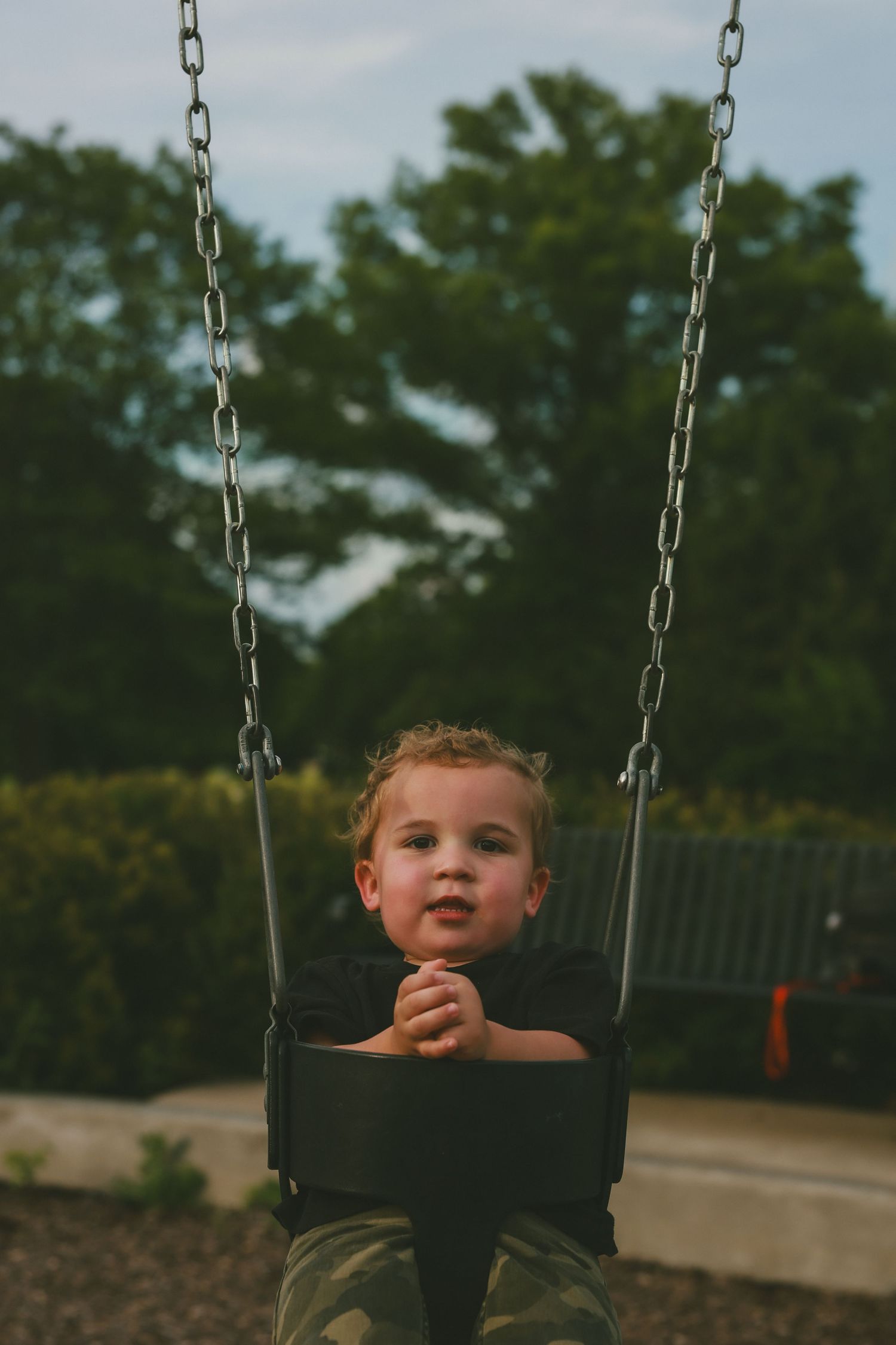 Child sitting in playground swing with curly hair against blurry green background with trees.