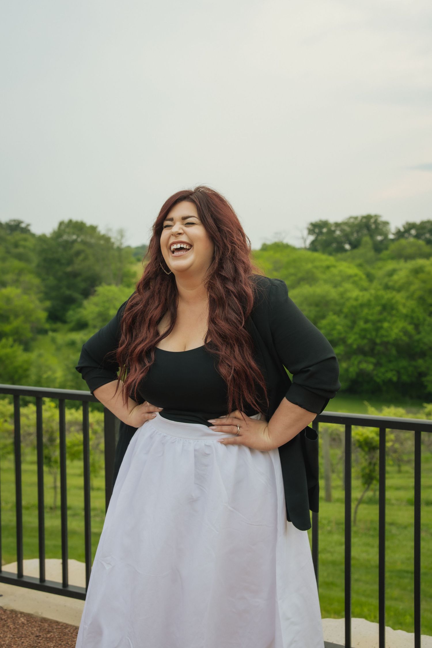 Woman in a black top and white tulle skirt poses joyfully on an outdoor balcony overlooking lush green scenery.