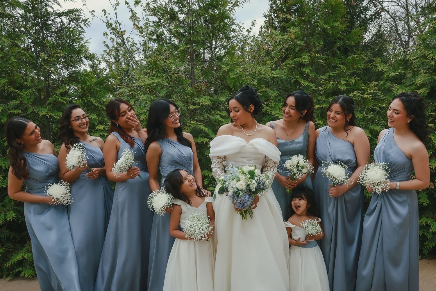 Wedding party in matching dusty blue bridesmaid dresses share a candid laughing moment with the bride in white.
