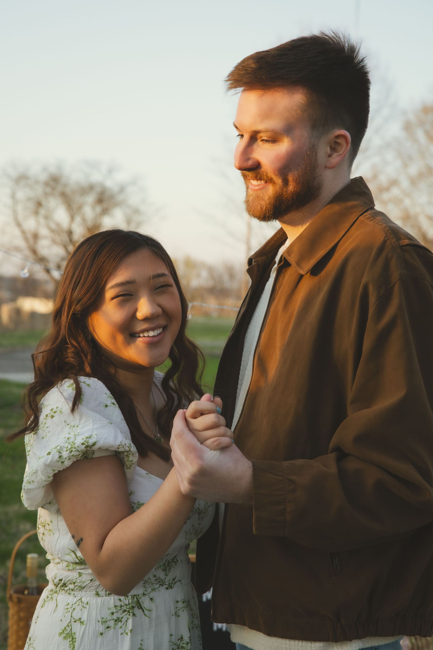 A couple laughs together outdoors during golden hour, with one wearing a brown jacket and the other in a floral dress.