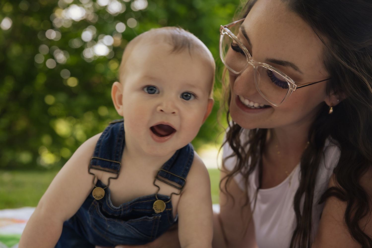 Parent and baby in overalls share joyful moment together on sunny day in park outdoor setting.