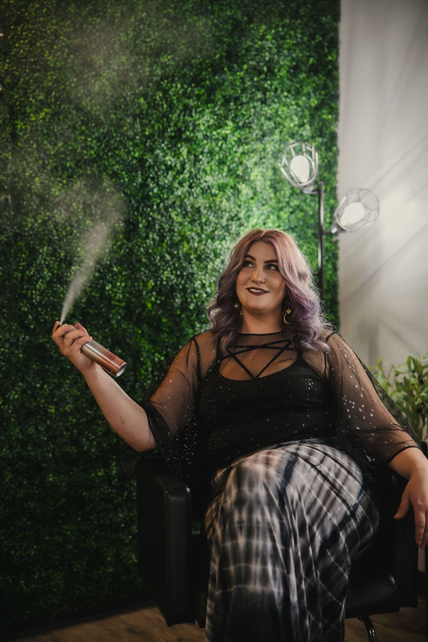 A person in a black mesh top and plaid skirt poses against a green vine wall with decorative lighting fixtures.