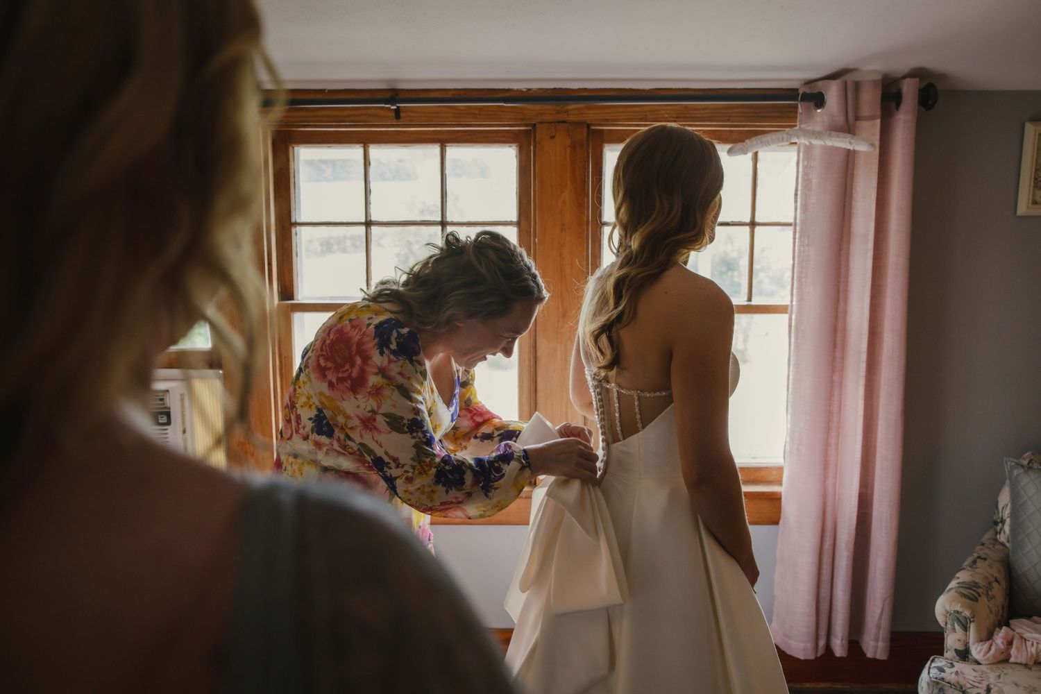 A tender moment as a bridal party helps adjust a wedding dress by a sunlit window during wedding preparations.