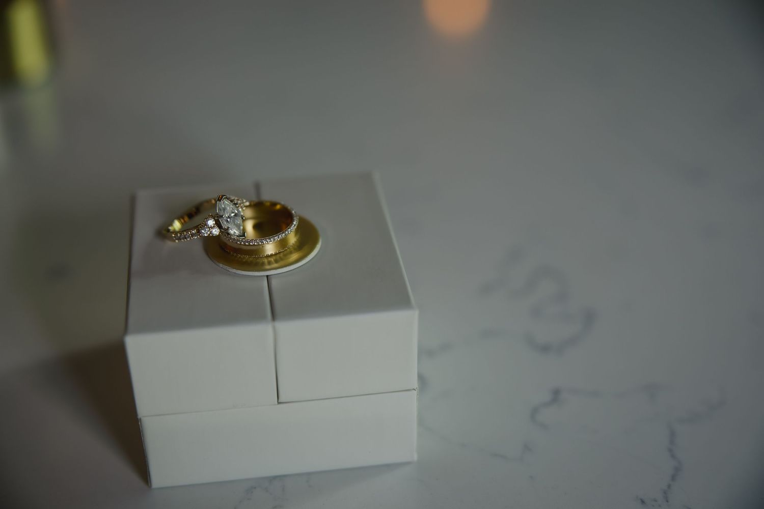 Gold engagement ring with diamond sits atop a white jewelry box against a light colored surface.