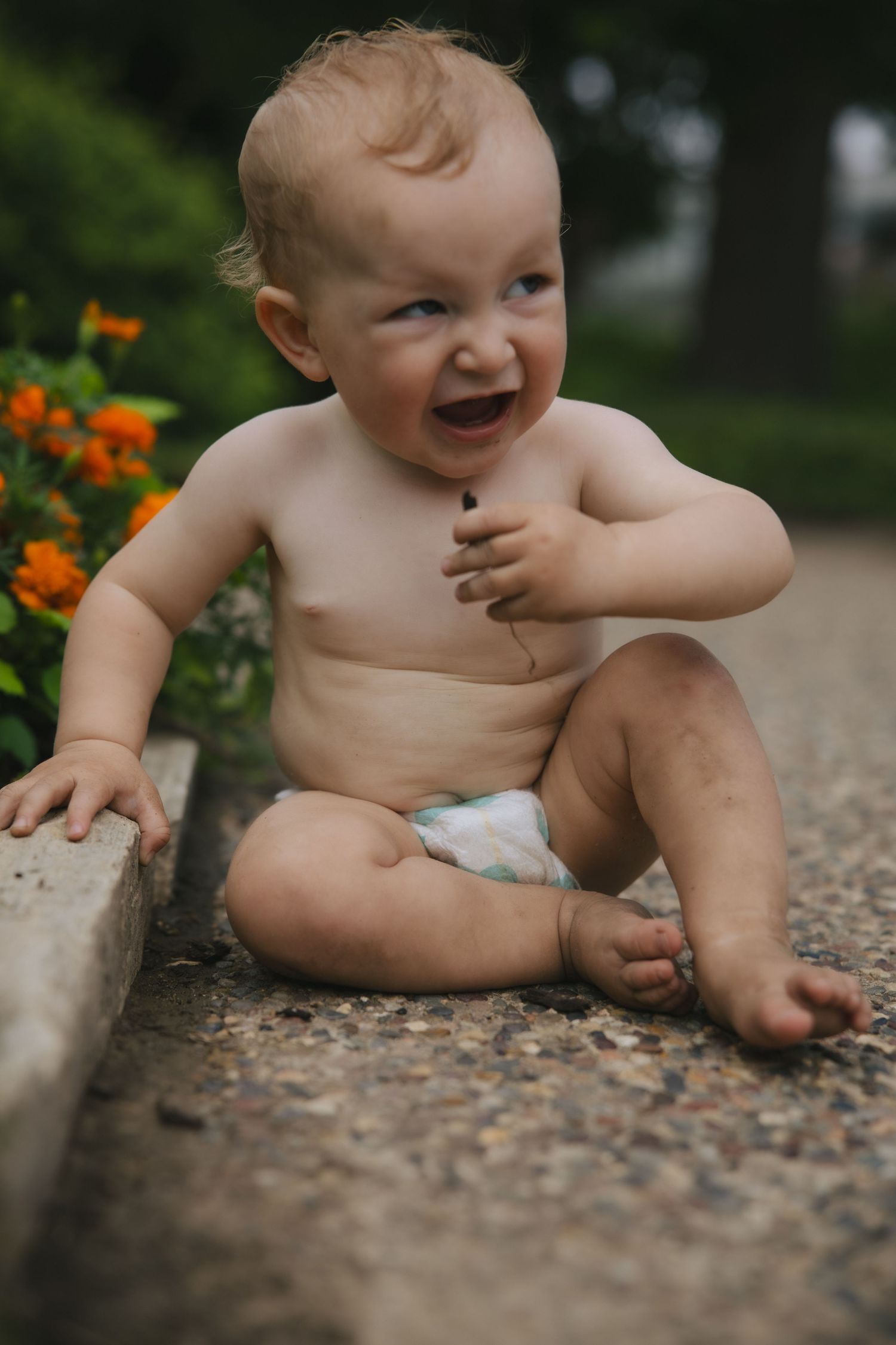 Happy baby wearing a diaper sits on a stone path next to vibrant orange flowers in a garden.