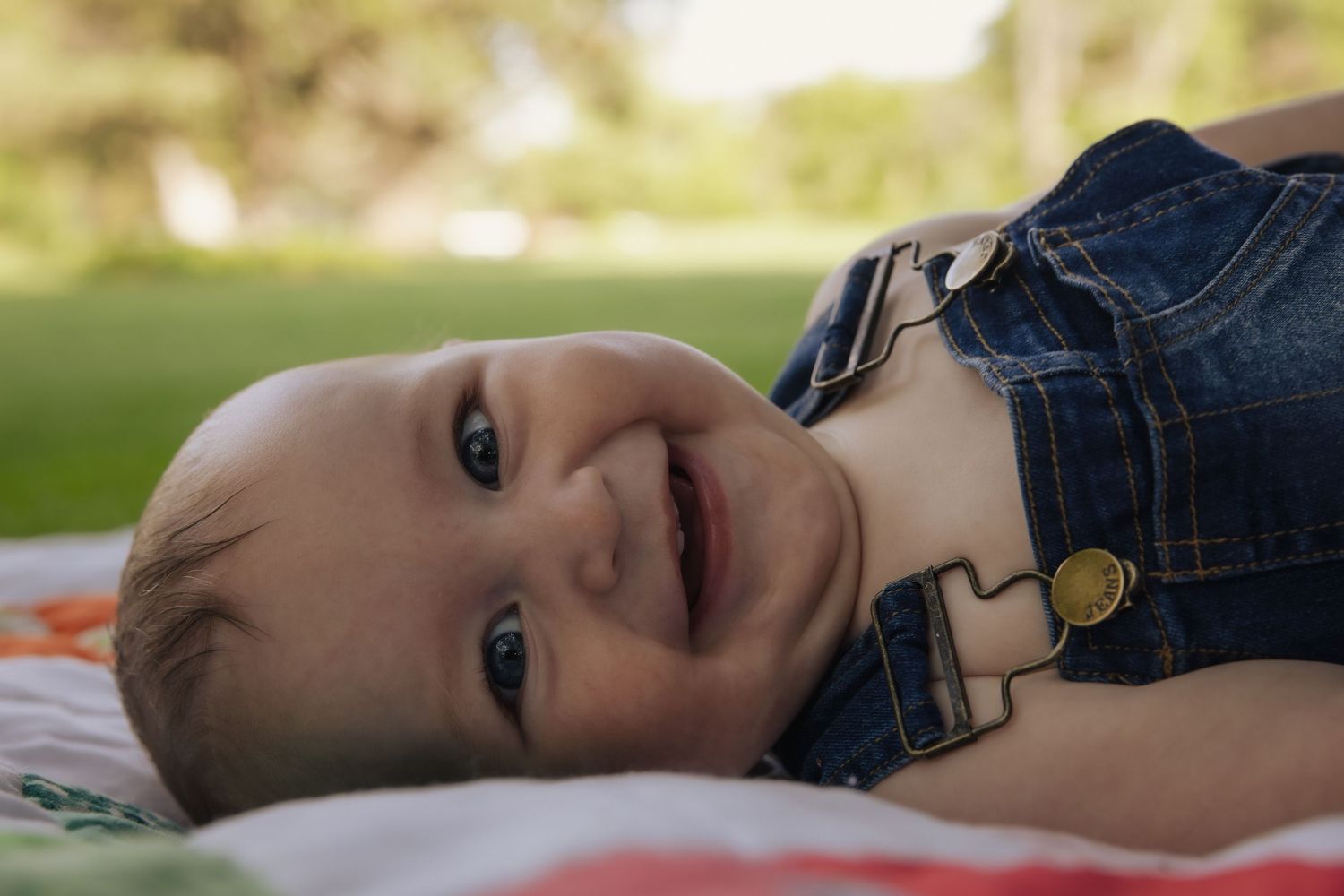 A young child in denim overalls lies on a blanket outdoors with a bright smile on their face.