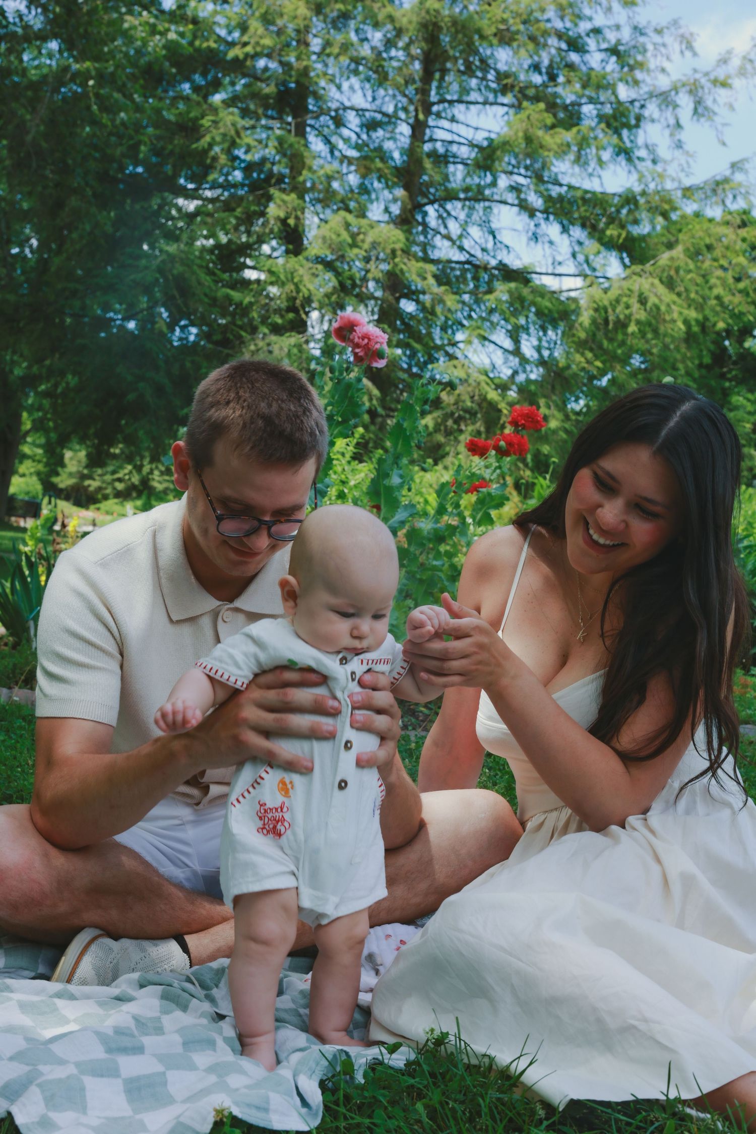 A family enjoys a summer day together on a picnic blanket in a lush green garden with red flowers in the background.